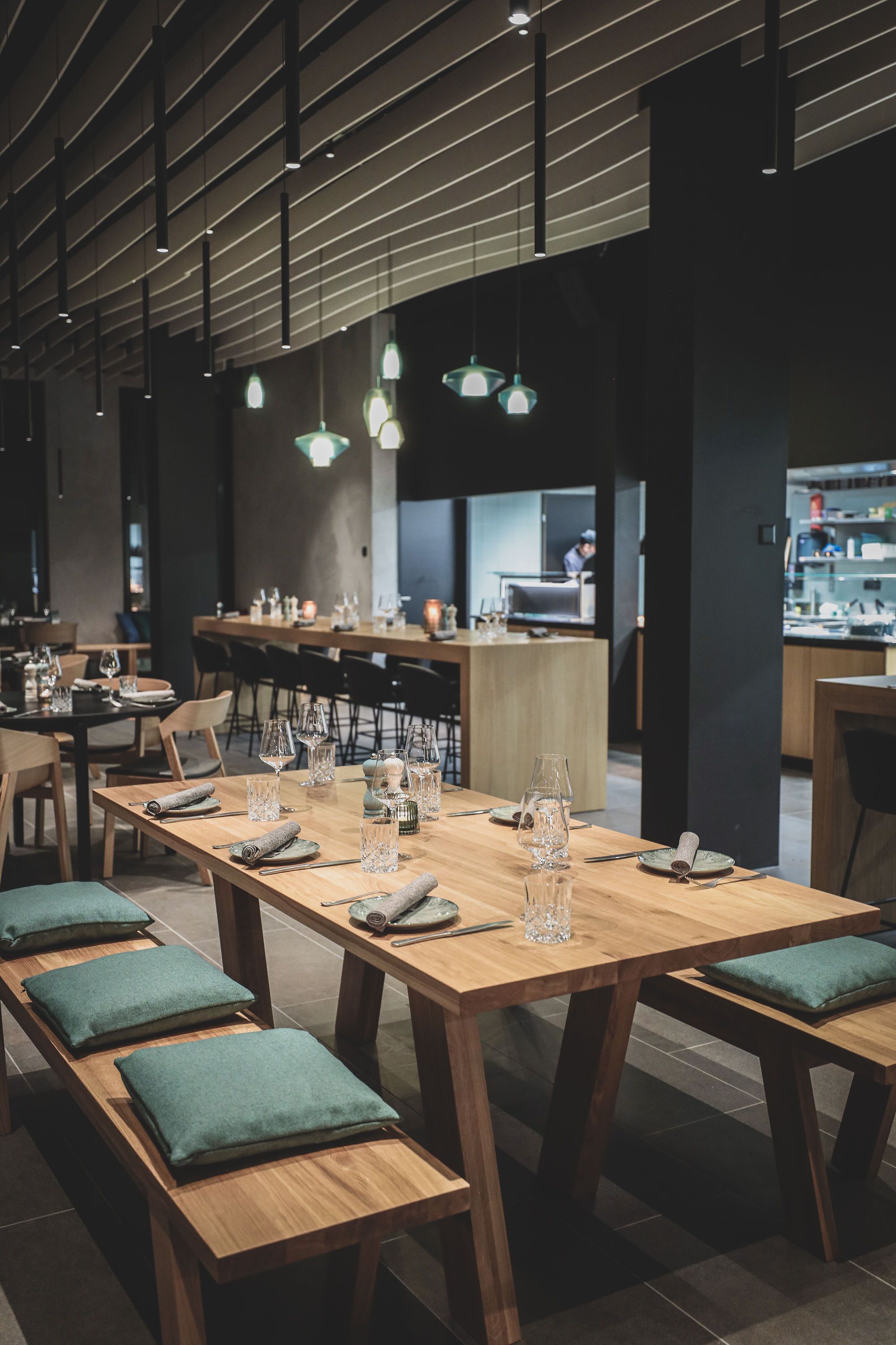 Un restaurante moderno con mesas y bancos de madera, una cocina al fondo e iluminación colgante.