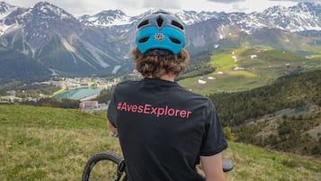 A person with a helmet and a black t-shirt with a hashtag, overlooking a mountainous landscape with a lake and buildings in the distance.
