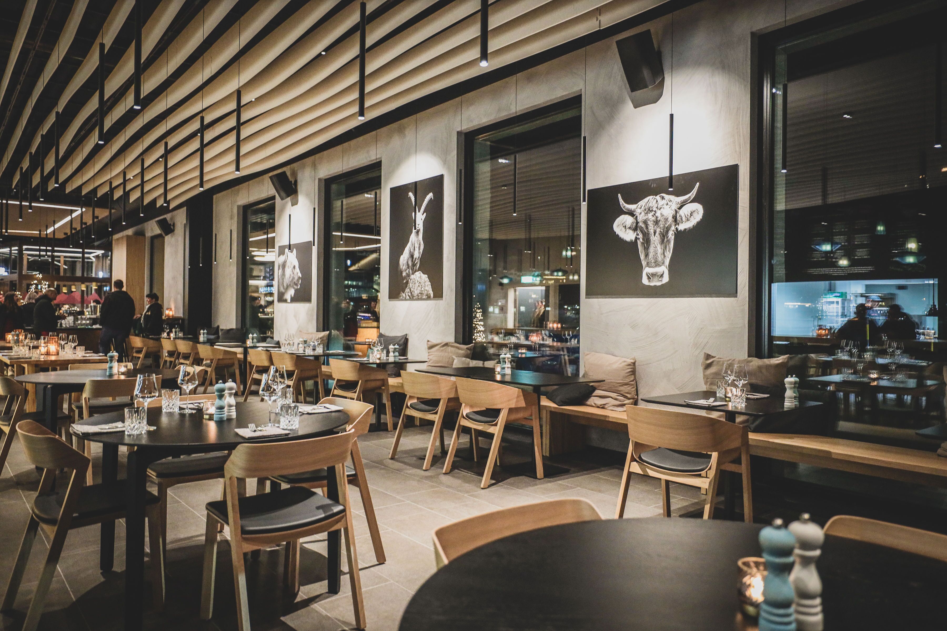 Un interior de restaurante moderno con mesas y sillas de madera, ventanas amplias y obras de arte en las paredes.