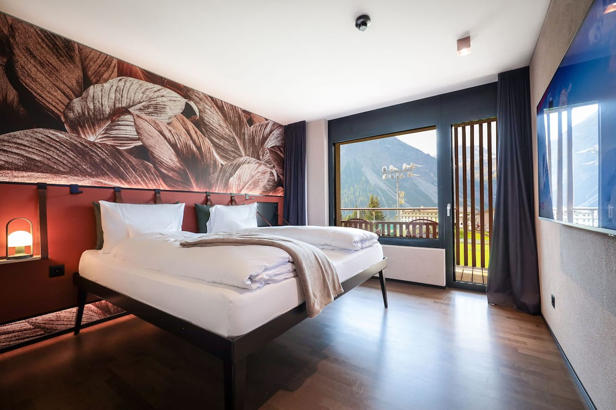 Una habitación de hotel moderna con una cama grande, vista a las montañas a través de la ventana y un mural en la pared.