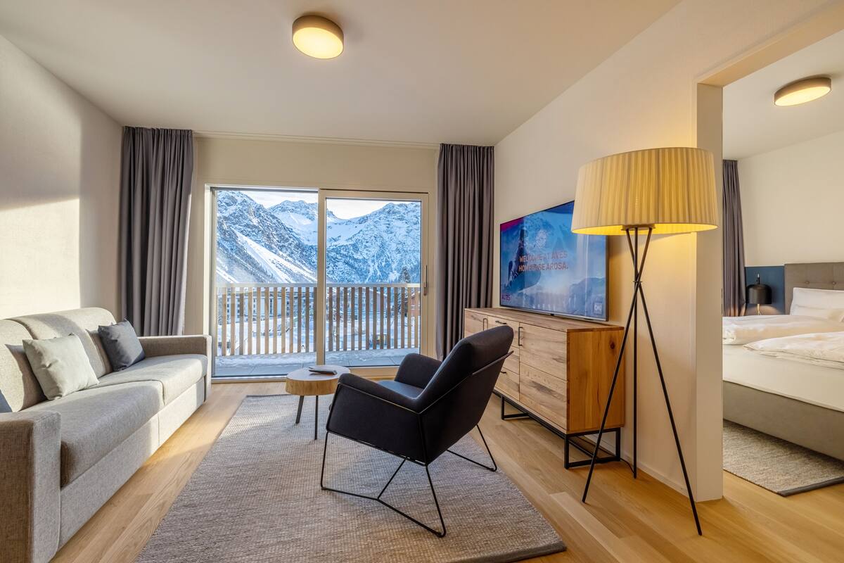 Una habitación de hotel moderna con sofá, sillón, escritorio, lámpara, televisor y cama.