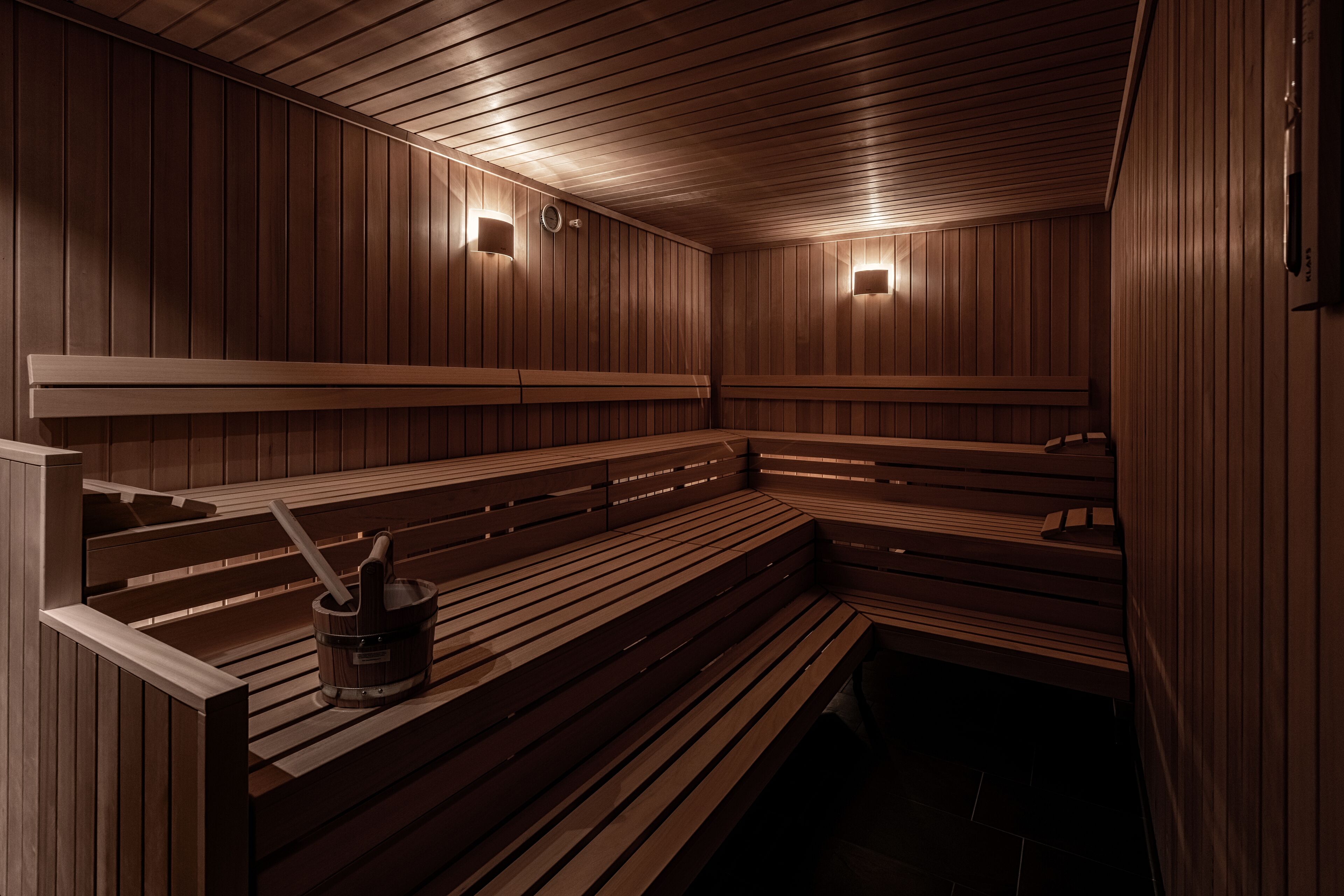 A sauna with wooden benches and walls, two wall-mounted lights, and a bucket with a brush.