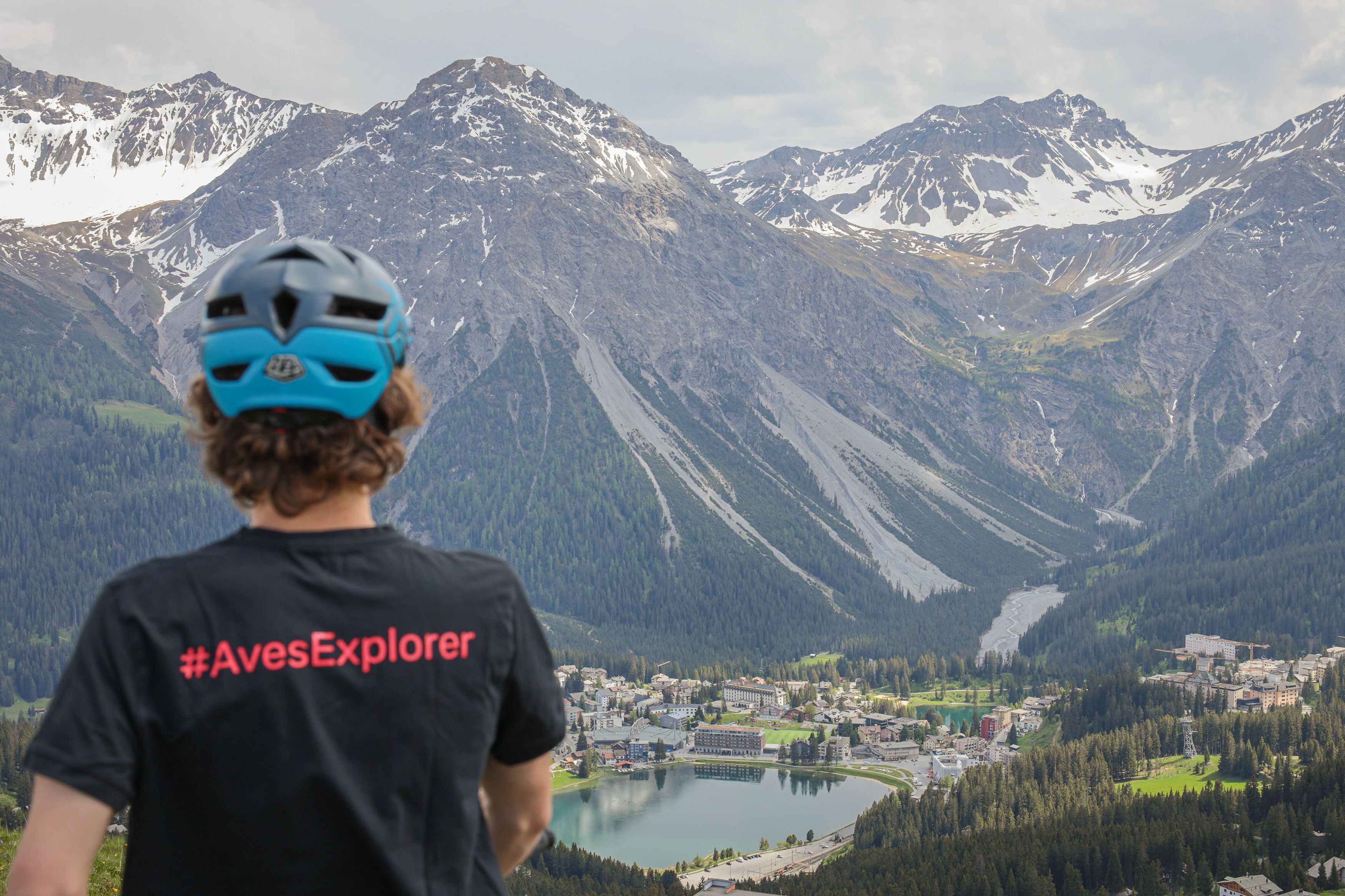 A person wearing a helmet and a black t-shirt with the text "#AvesExplorer" standing in front of a mountainous landscape with a lake and buildings in the valley.