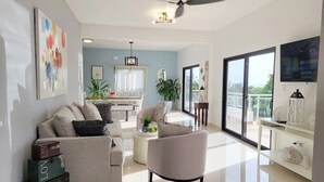 A modern living room with a sofa, armchair, and a dining area with a table and chairs. There is a balcony with a railing and a view of the outdoors. - Luxury Apt w/ Pool 5-min Walk to Beach & Ocean World (Cofresi)