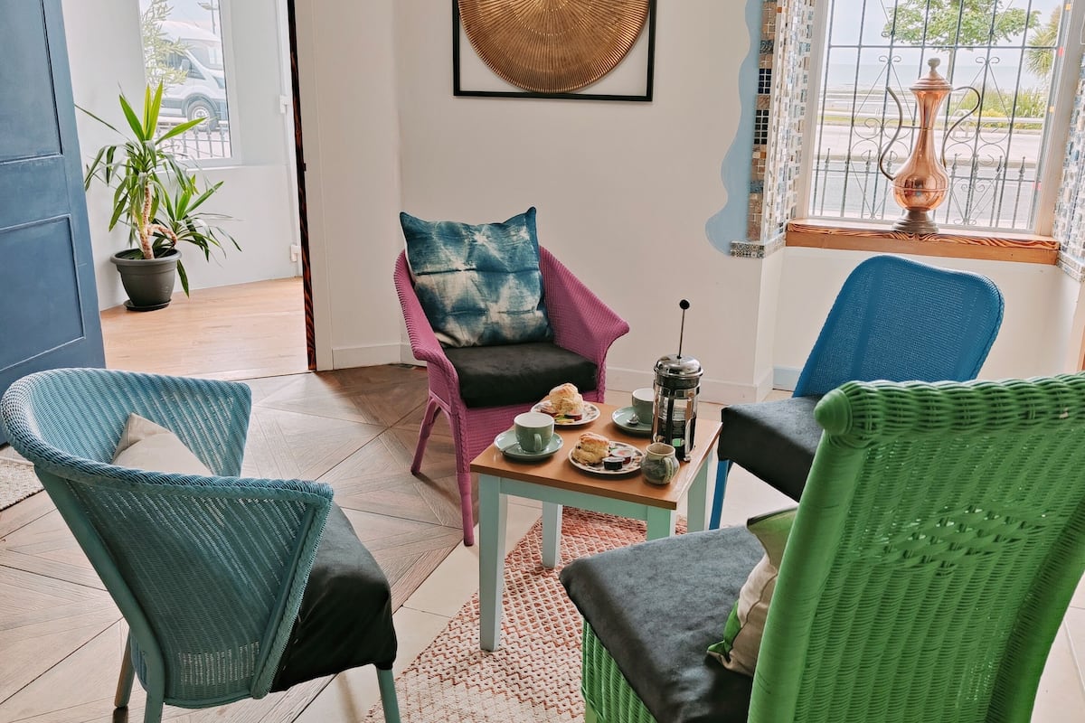 A cozy room with wicker chairs, a coffee table with pastries, and a view through a window with a decorative vase.