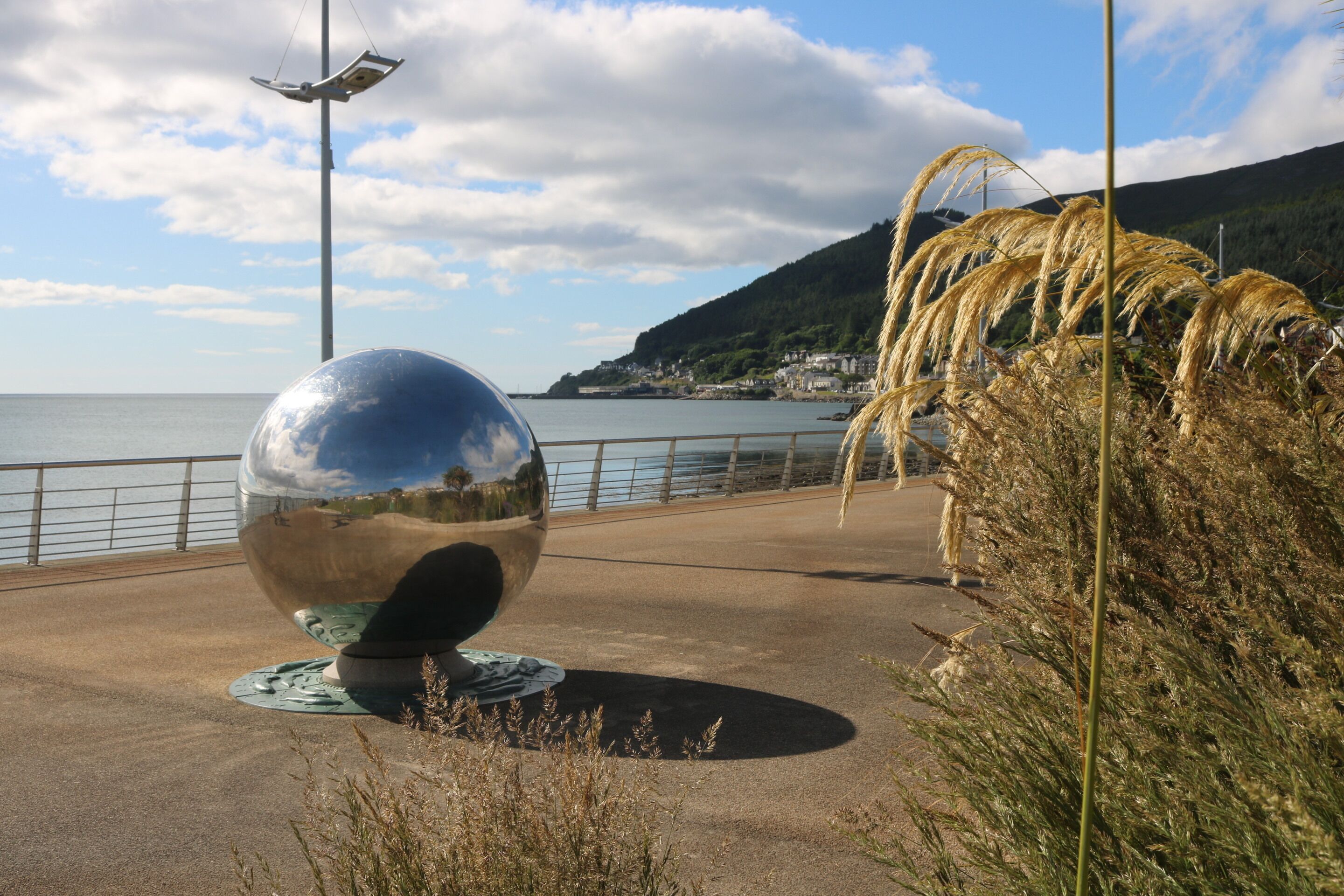 Una escultura reflectante en un camino pavimentado junto al mar, con barandilla y vegetación costera.