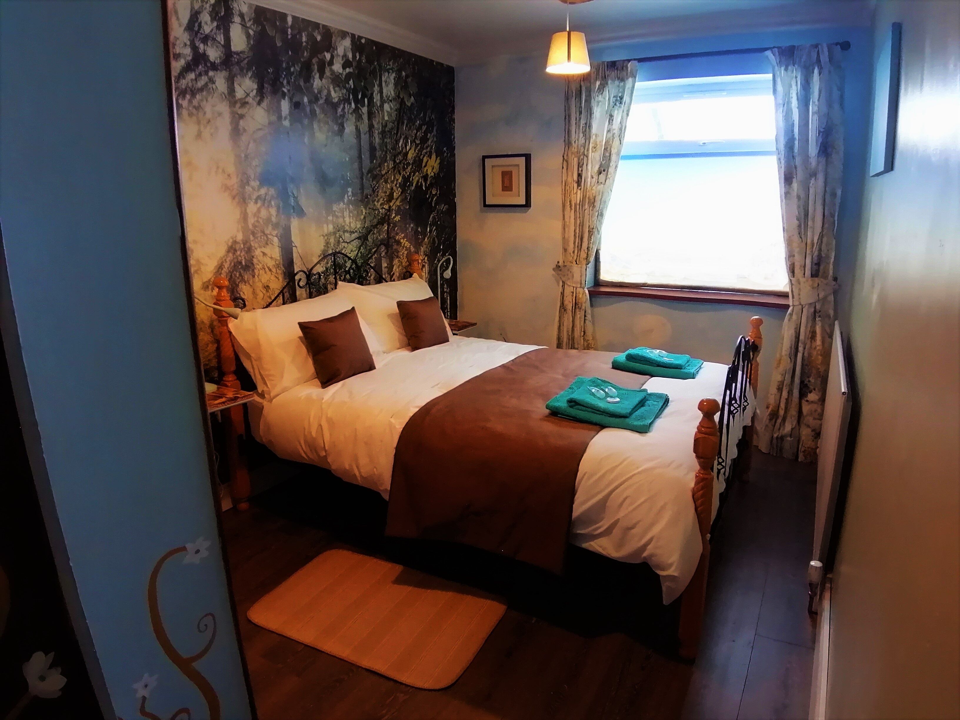 Un dormitorio con cama, una ventana con cortinas, un cuadro en la pared y una lámpara de noche.