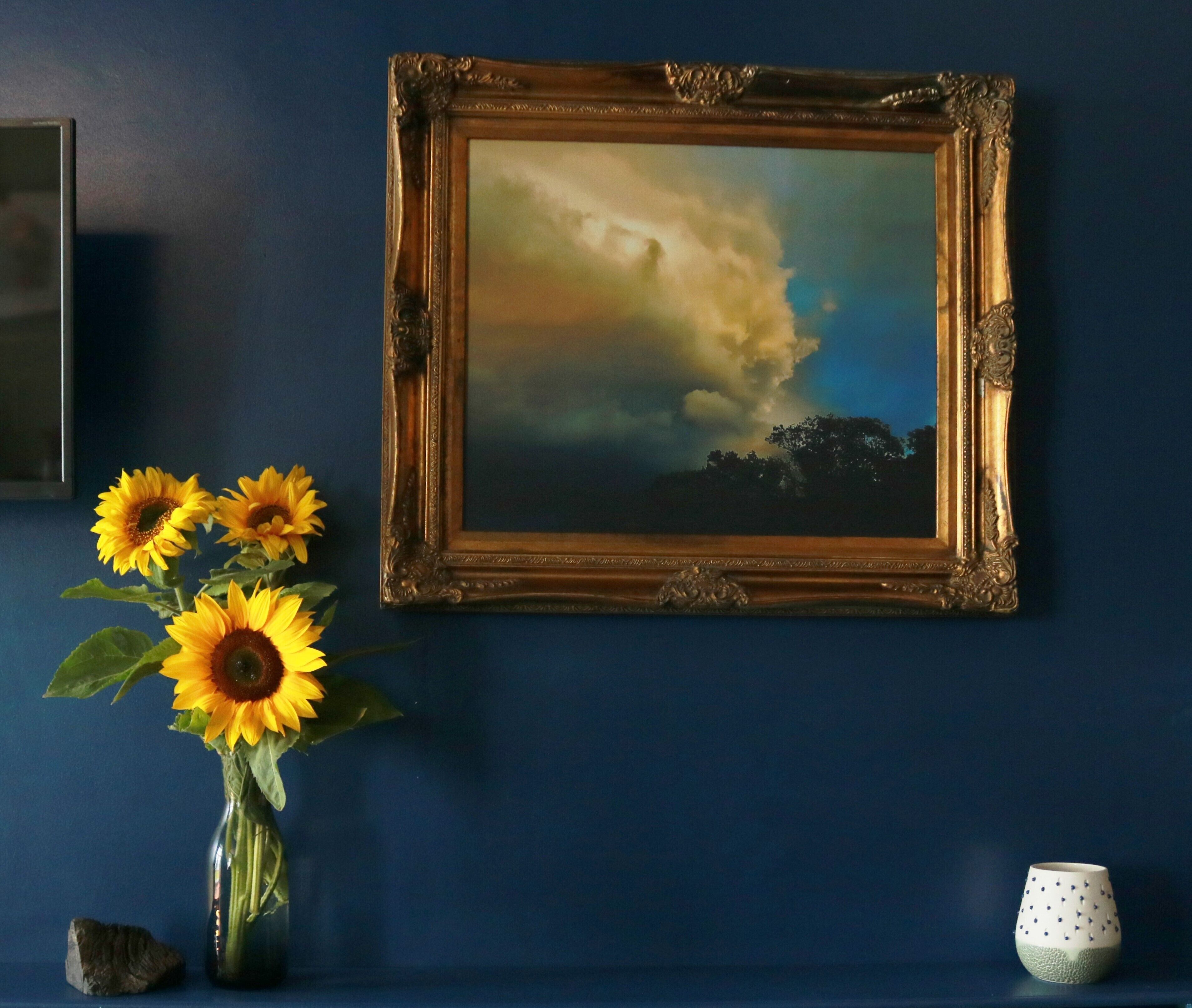 A framed painting of a cloudy sky on a blue wall, a vase with sunflowers, and a small decorative cup.