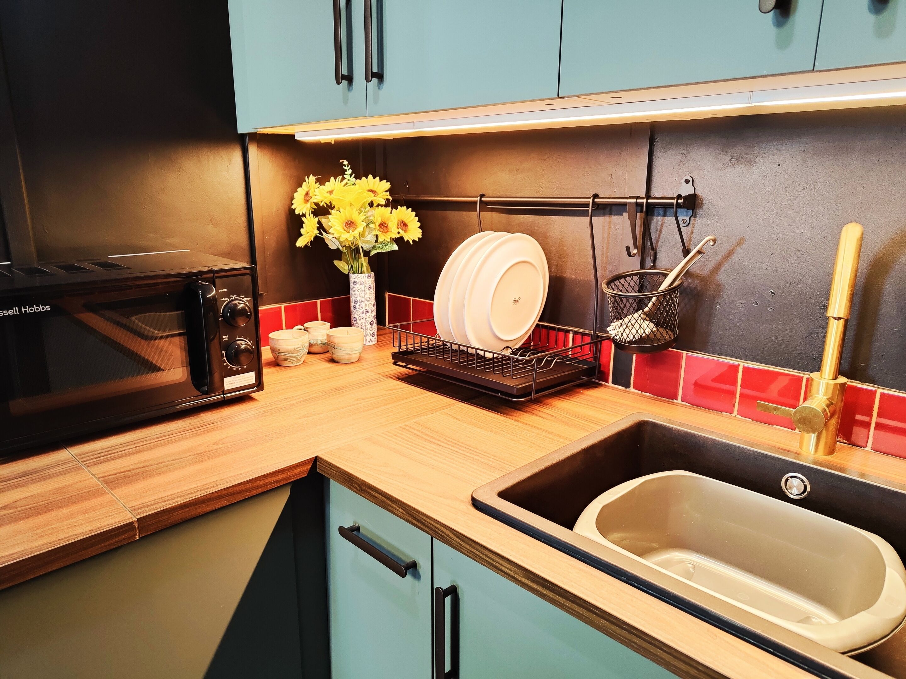 A kitchen with a black microwave, a dish rack, a sink, and a vase with flowers.