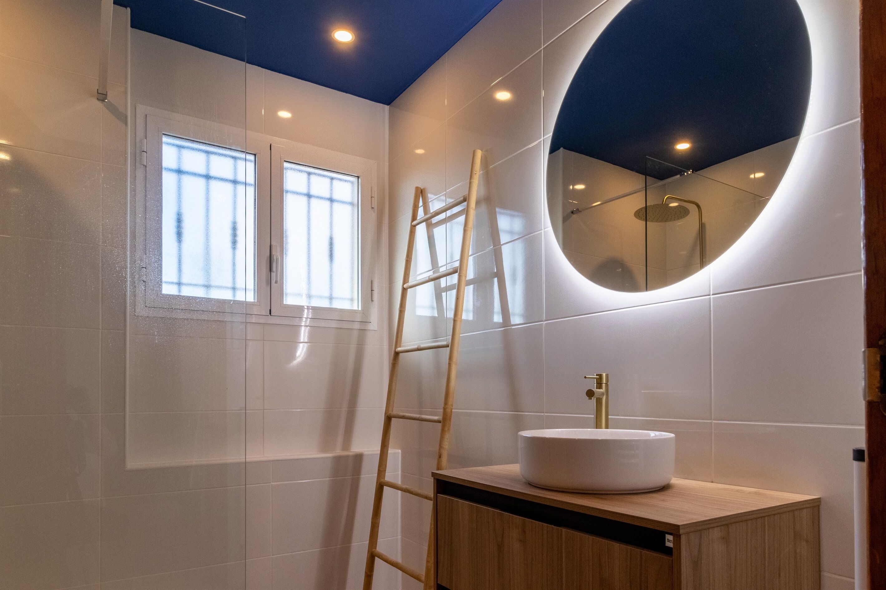 A modern bathroom with a white tiled wall, a round mirror, a wooden ladder, and a white sink.