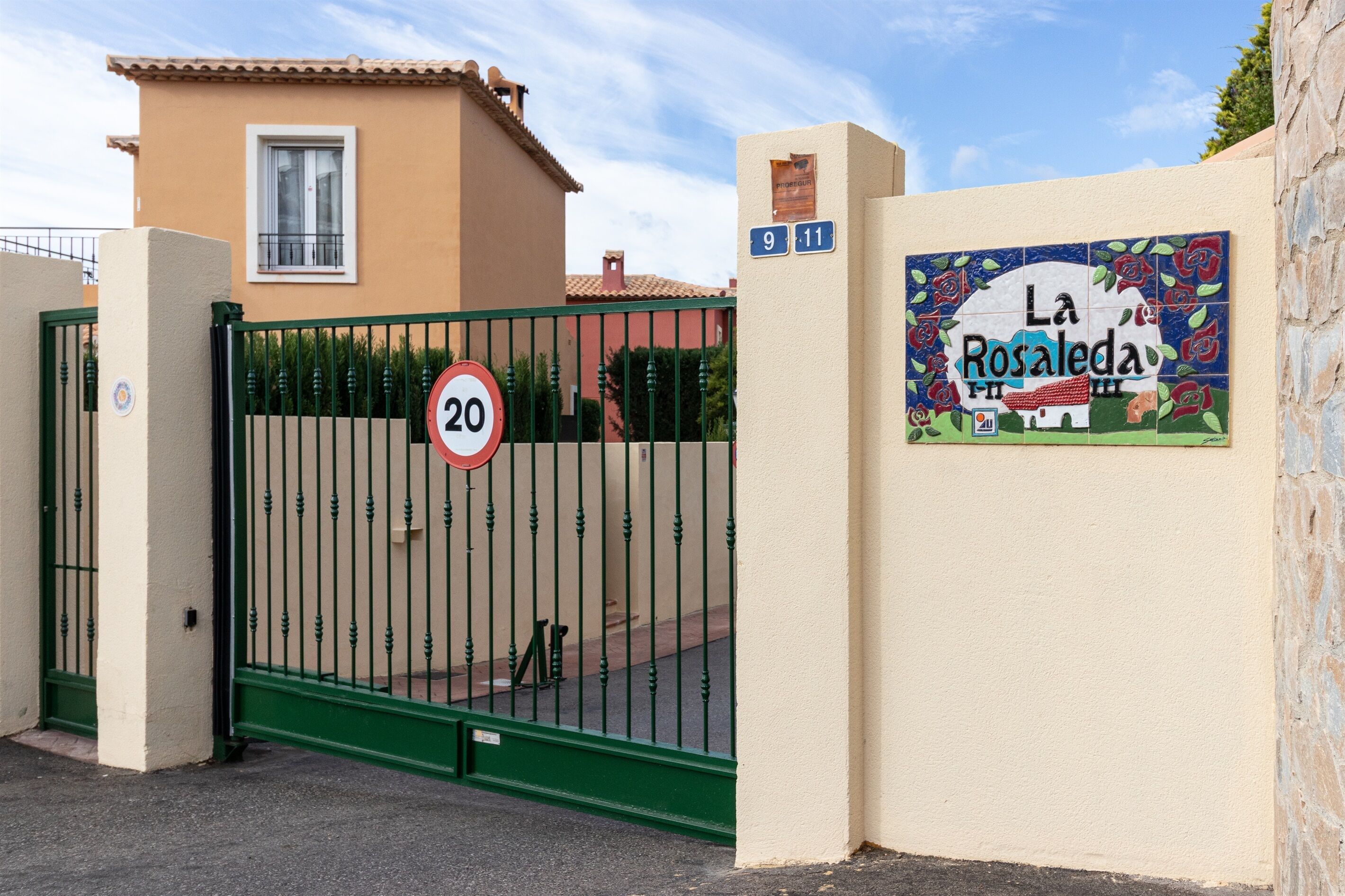 Una entrada tancada amb un senyal de límit de velocitat de '20', una casa amb un balcó i un cartell de colors que diu 'La Rosaleda II'.