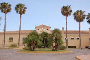 A building with a beige facade, a central entrance with a triangular pediment, and palm trees surrounding the entrance.