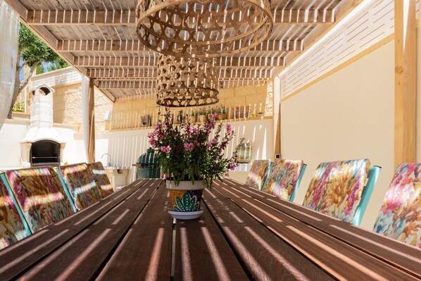 An outdoor dining area with a wooden table and chairs, a hanging light fixture, and a potted plant with flowers.