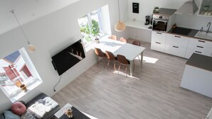 Una zona d'estar i menjador moderna i diàfana, amb cuina, sofà, taula de dinar i televisió penjada a la paret.
