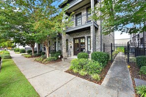 A two-story residential building with a stone facade, a balcony on the upper floor, and a well-maintained front yard with trees and shrubs. - Bama Getaway - Walk to Downtown & Campus!  Luxurious and cozy furnishings! (Tuscaloosa)