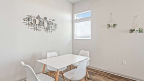 Une pièce moderne avec une table et des chaises blanches, une œuvre d’art géométrique accrochée au mur, une fenêtre avec des stores et une petite plante suspendue.