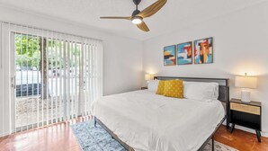 Un dormitorio con cama, ventilador de techo, mesita de noche con lámpara y ventana con persianas.