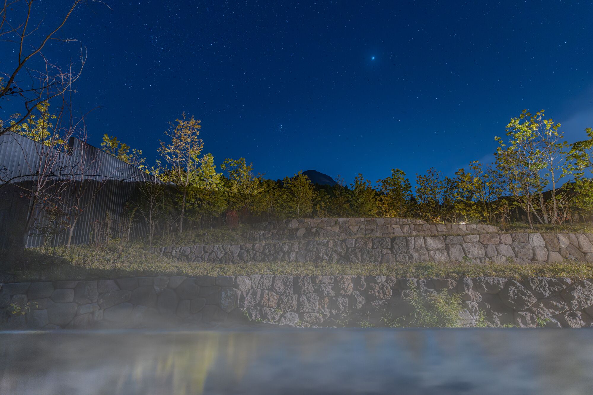 A stone wall with a reflection in a body of water, trees, and a mountain in the background under a starry night sky.