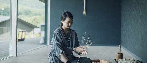 A person sitting on the floor in a room with a large window, holding a branch. There are boxes and a wooden object on the floor.