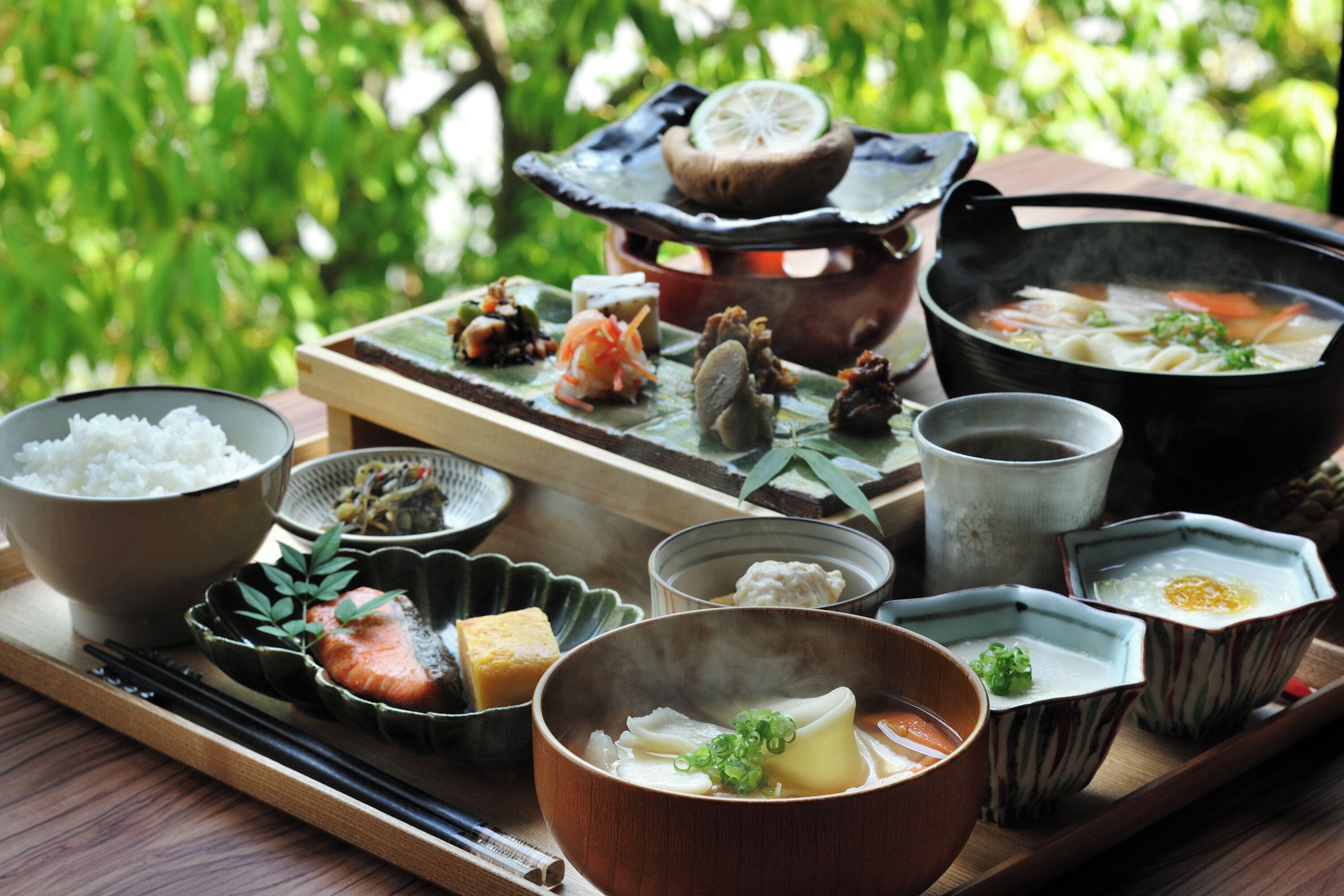 A variety of Japanese dishes on a wooden tray, including rice, salmon, and soup, set against a backdrop of greenery.