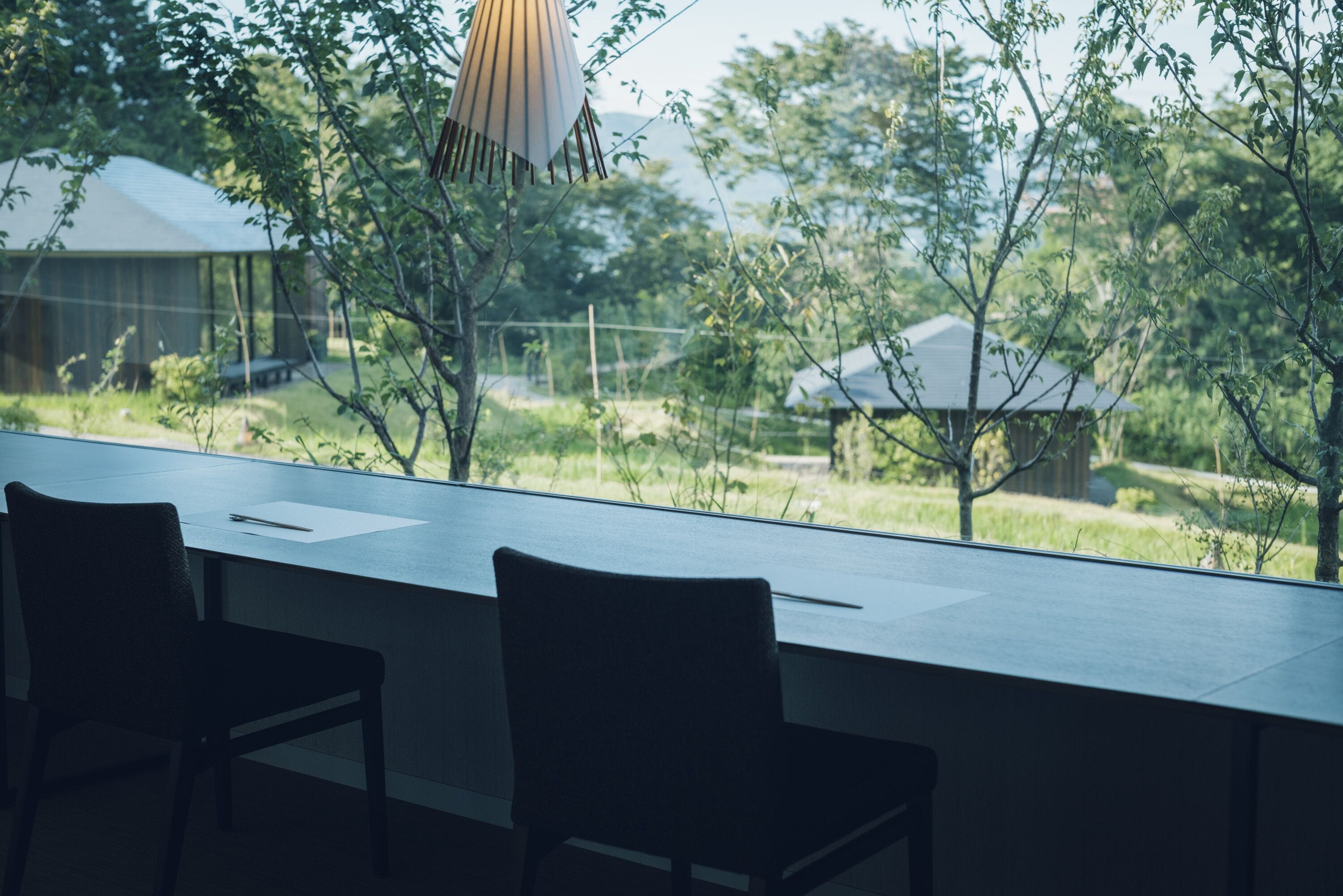A modern room with a large window overlooking a garden and a building. There are two chairs and a table in the room.