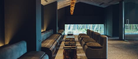 A modern interior with a long row of benches and a central coffee table, large windows, and a ceiling with a unique light fixture.