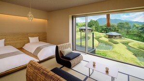 Et moderne hotelværelse med to senge, en sofa, et bord og et stort vindue med udsigt over terrasserede marker og en bygning i det fjerne.