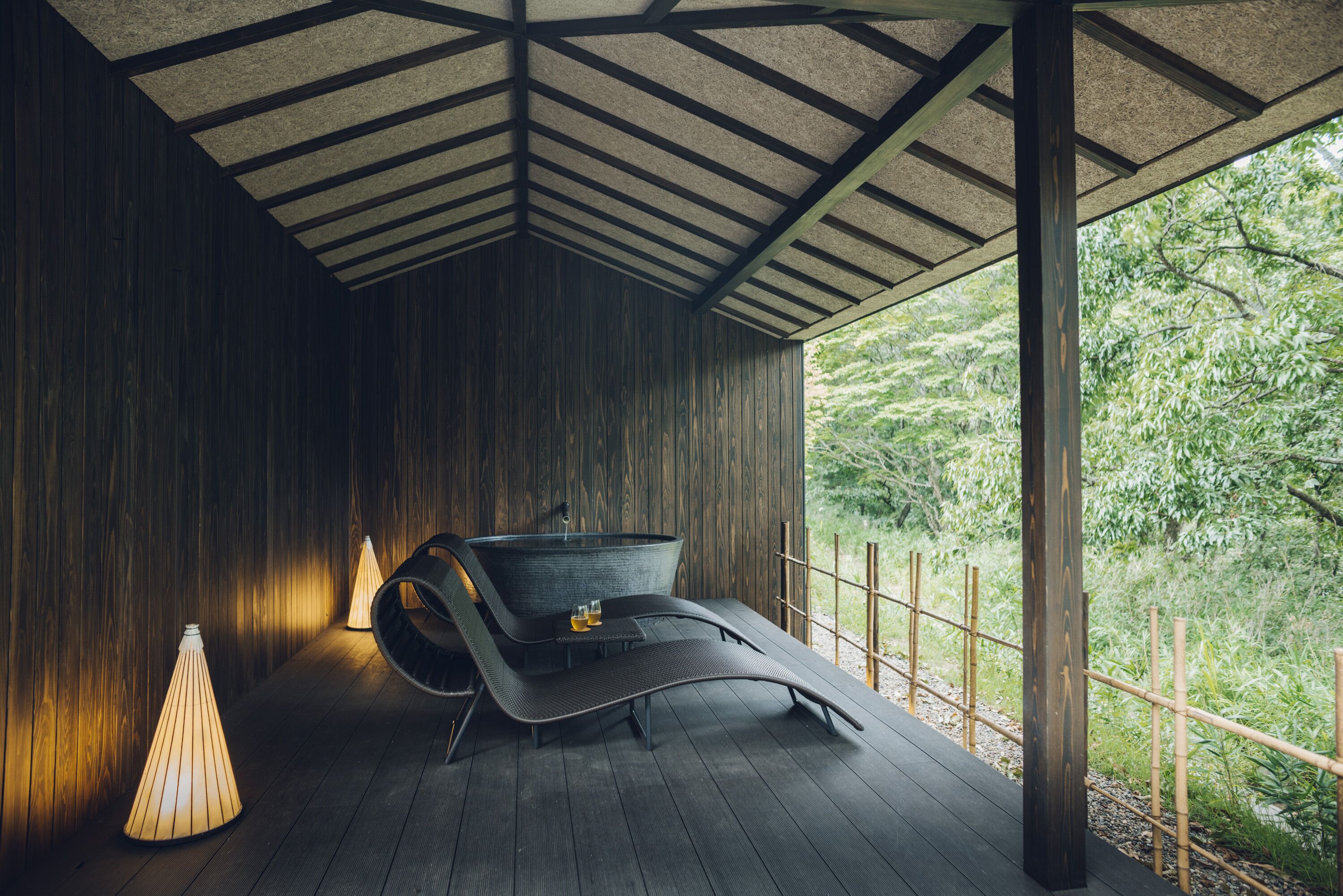 A modern, dark wooden room with a curved bench and a small table, a hanging light fixture, and a view of greenery outside.