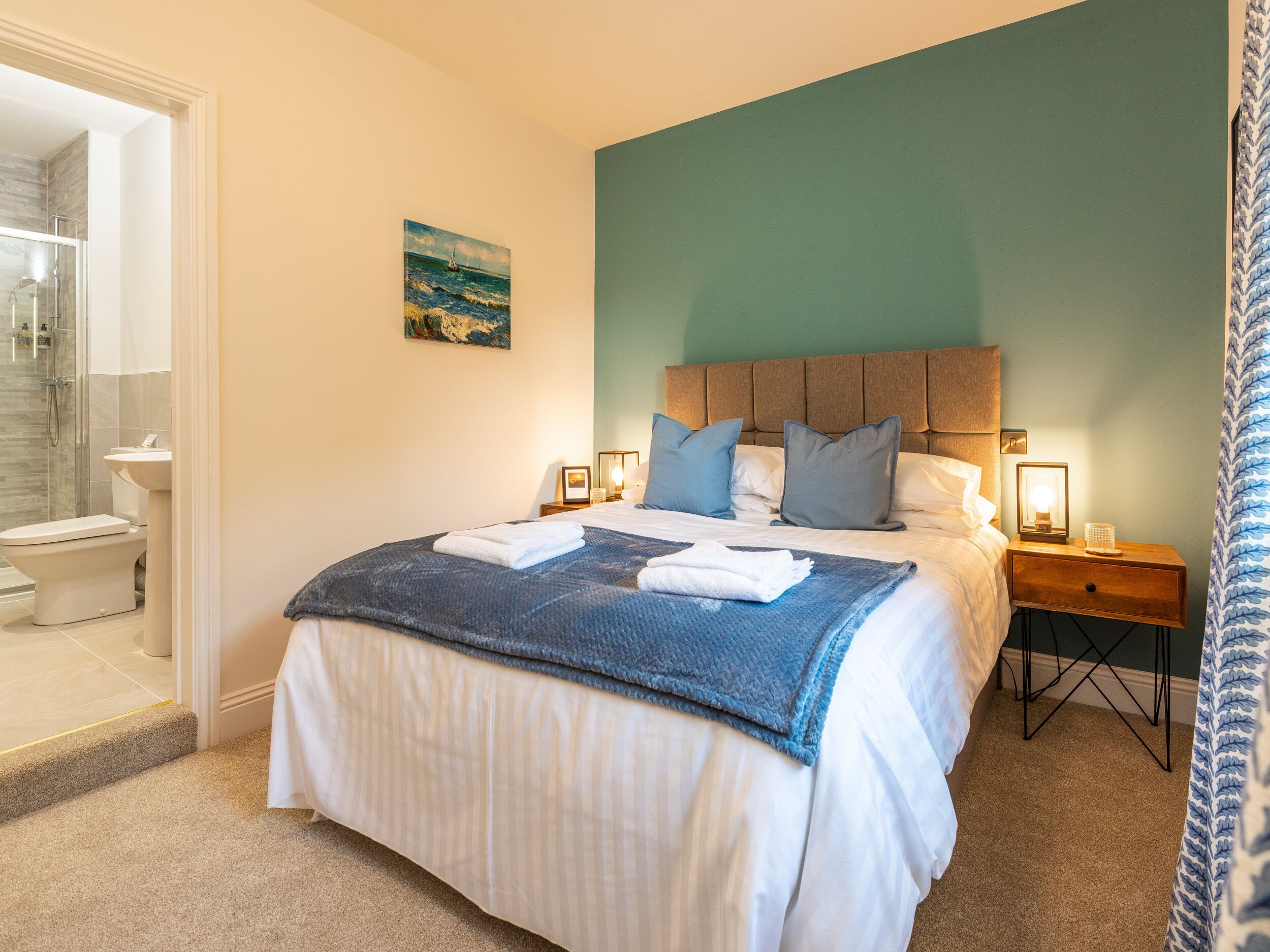 Une chambre à coucher avec un lit, une table de chevet, un tableau et une salle de bain visible à travers une porte ouverte.