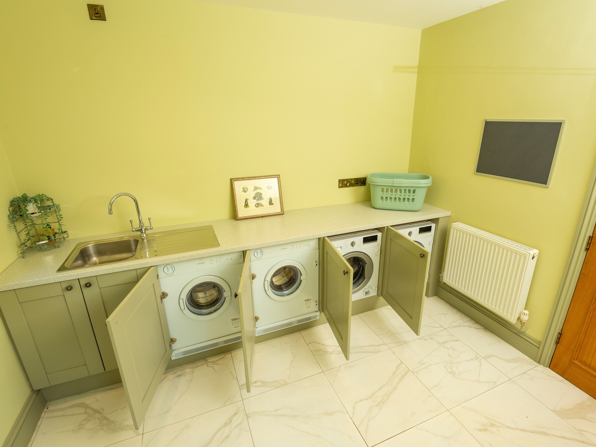 A compact kitchen with a sink, two washing machines, a framed picture, and a radiator.
