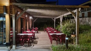 Un área de comedor al aire libre con mesas y sillas de madera, un paseo techado y un muro de piedra al fondo.