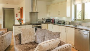 A modern kitchen with white cabinets, stainless steel appliances, and a built-in oven. There is a dining area with a sofa and cushions. - The Mews (Newton Abbot)