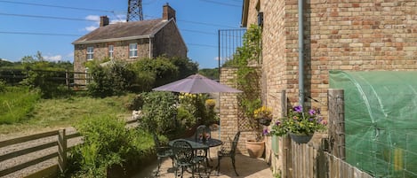 A stone house with a patio, a table, and chairs. There is a wooden fence and a green netting structure.