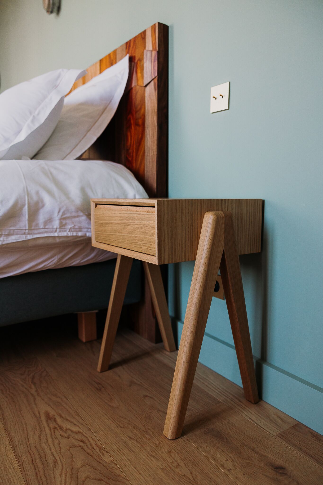 Une table de chevet en bois avec un tiroir, un lit recouvert de draps blancs et un mur bleu clair.