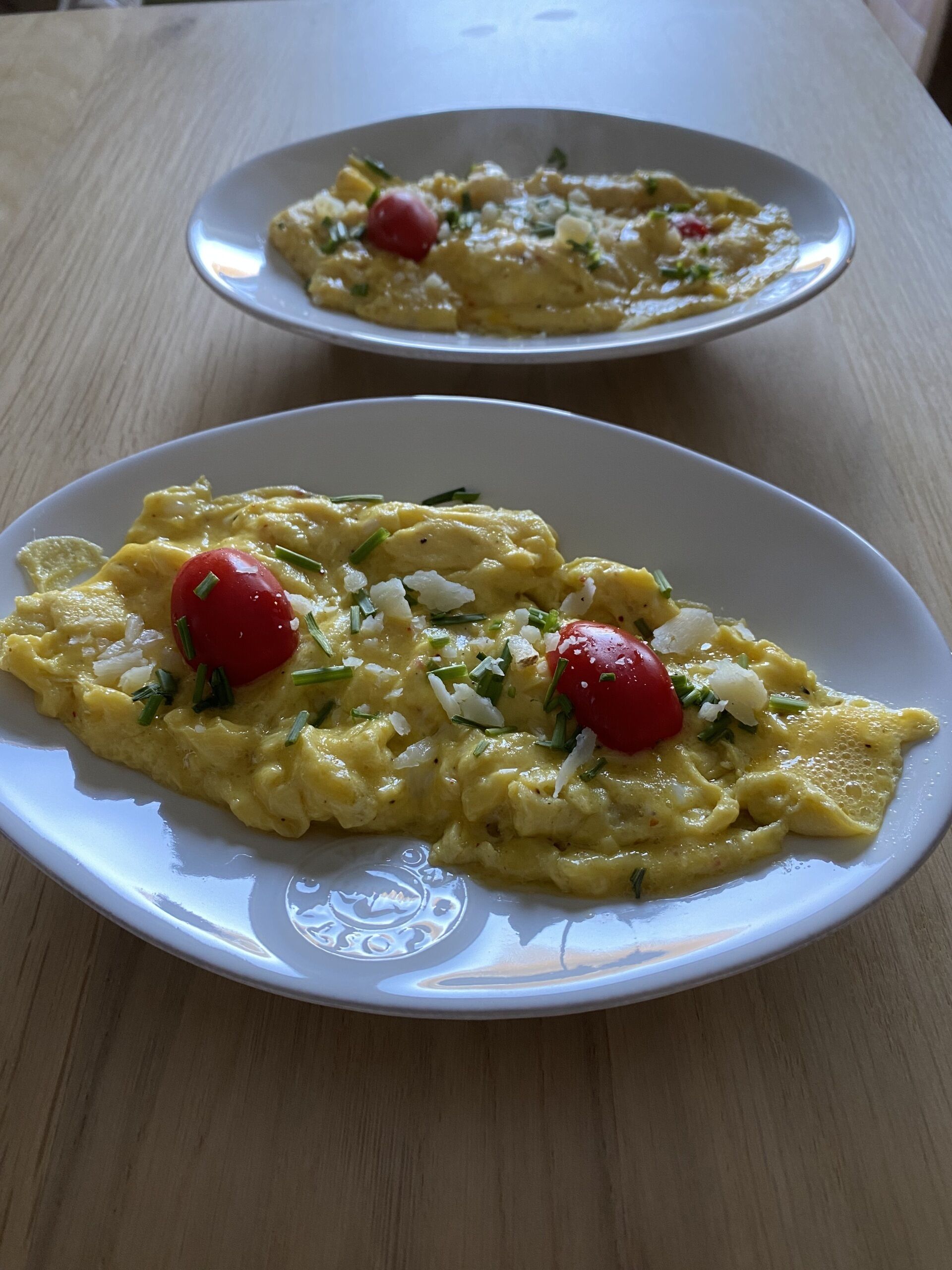 Dos platos blancos con huevos revueltos adornados con cebollín y tomates cherry sobre una mesa de madera.