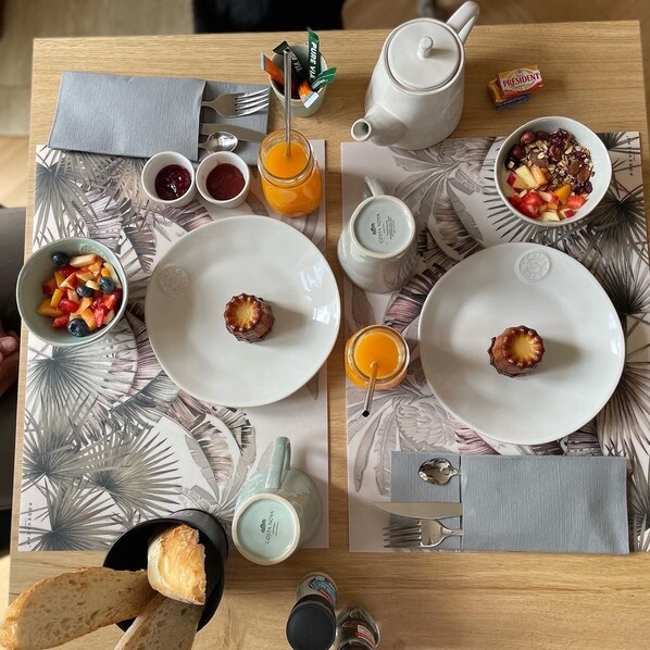 Una mesa de desayuno con platos, una tetera, un tarro de miel, un bol de fruta y una caja de cereal President.