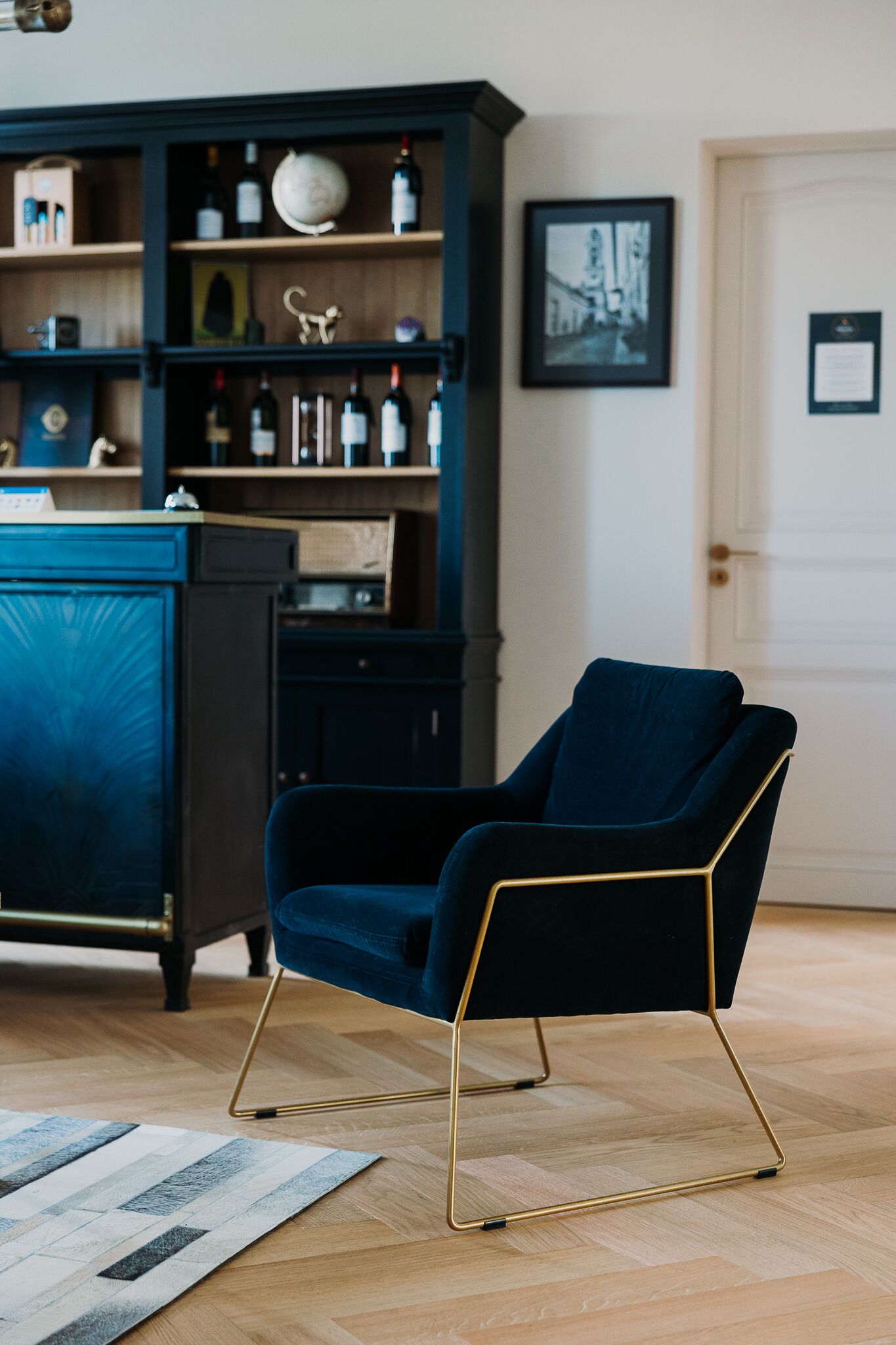 A room with a dark blue armchair with gold legs, a wooden floor, a blue cabinet with various items, and a framed picture on the wall.