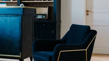 A room with a dark blue armchair with gold legs, a wooden floor, a blue cabinet with various items, and a framed picture on the wall.
