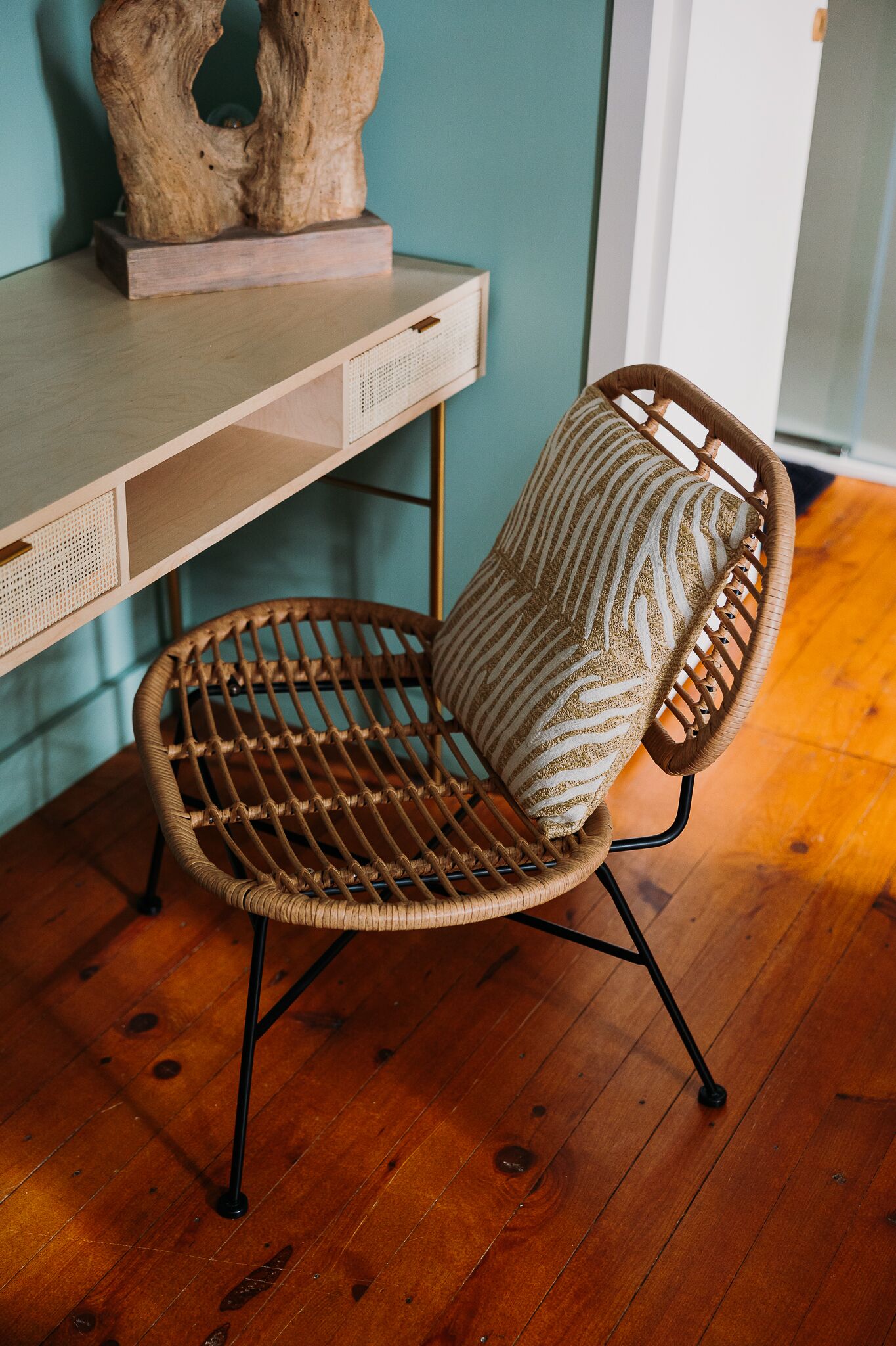 Una silla de mimbre con un cojín estampado, una escultura de madera sobre un escritorio y una pared color turquesa.