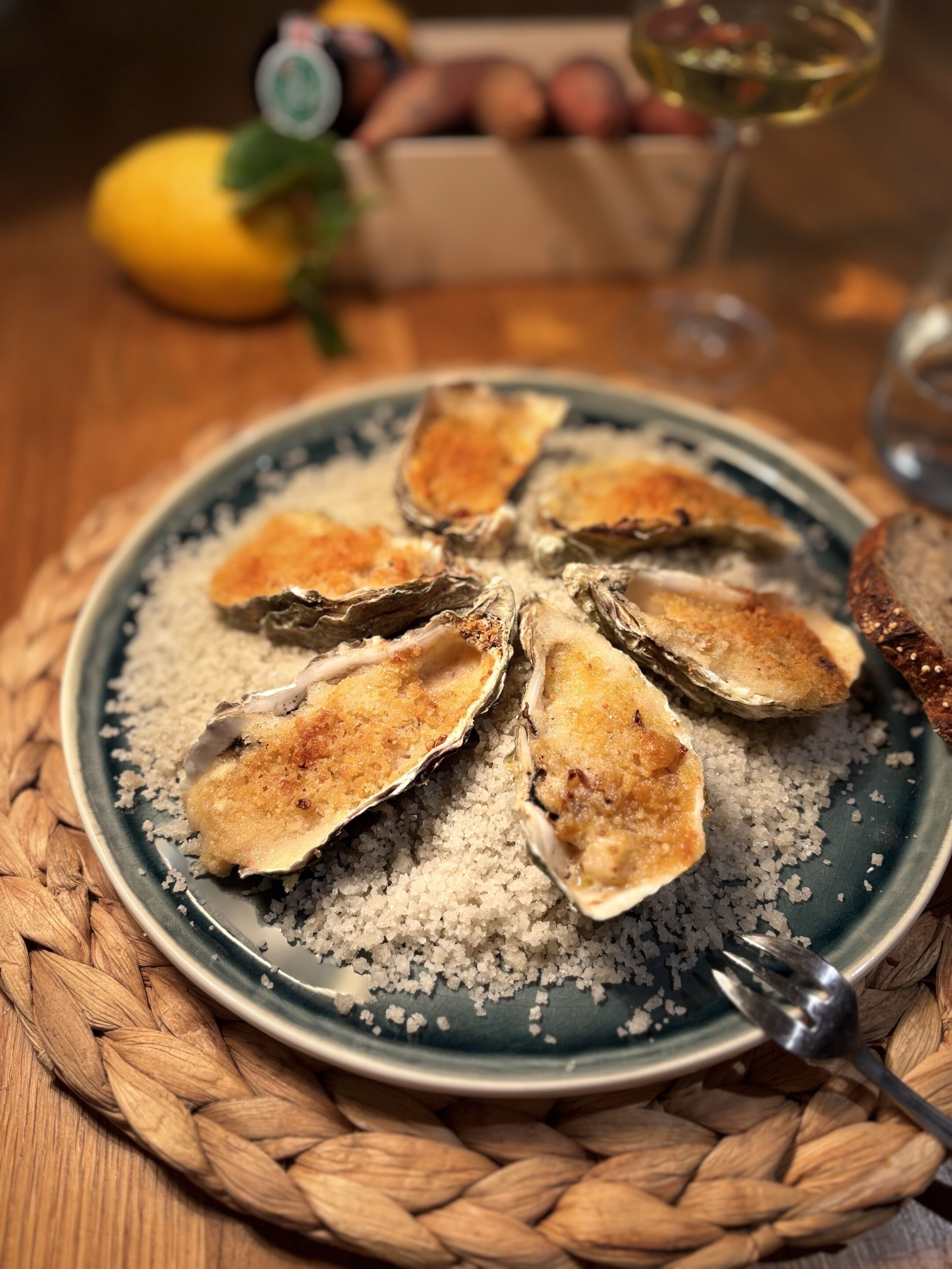 Een bord met oesters op hun schaal, bestrooid met grof zout, een vork en een glas witte wijn op een geweven mat.
