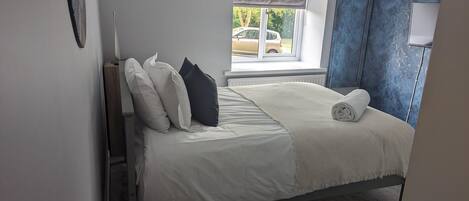 A neatly made bed with white and dark pillows, a rolled towel, and a window with a view of a car outside.