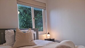 Una cama bien tendida con almohadas y toallas enrolladas, una lámpara de noche y una ventana con vistas a árboles.