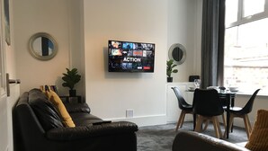 A living room with a black leather sofa, a smaller sofa with a yellow throw, a TV mounted on the wall, a dining table with chairs, and a window with curtains. - Bright Spacious 3-bed With Office & Patio, Preston (Preston)