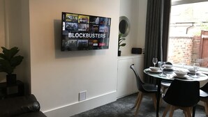 A living room with a flat-screen TV displaying a movie selection, a dining area with a table set for two, a potted plant, and a window with curtains. - Bright Spacious 3-bed With Office & Patio, Preston (Preston)