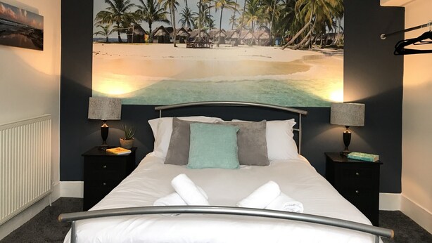 A bedroom with a bed, two bedside tables, a nightstand, a wall-mounted lamp, and a tropical beach mural. - Bright Spacious 3-bed With Office & Patio, Preston (Preston)