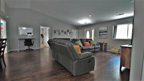 En rummelig stue med en grå hjørnesofa, et spiseområde med bord og stole, samt et køkkenområde med bordplade og hårde hvidevarer.
