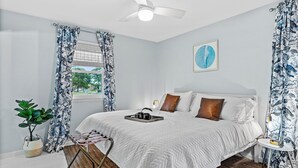 Une chambre à coucher avec un lit, un ventilateur de plafond, une fenêtre avec des rideaux, une plante et un tableau encadré au mur.