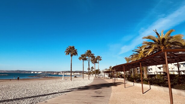 Une promenade en bord de mer avec une passerelle en bois, des palmiers et un ciel bleu azur.