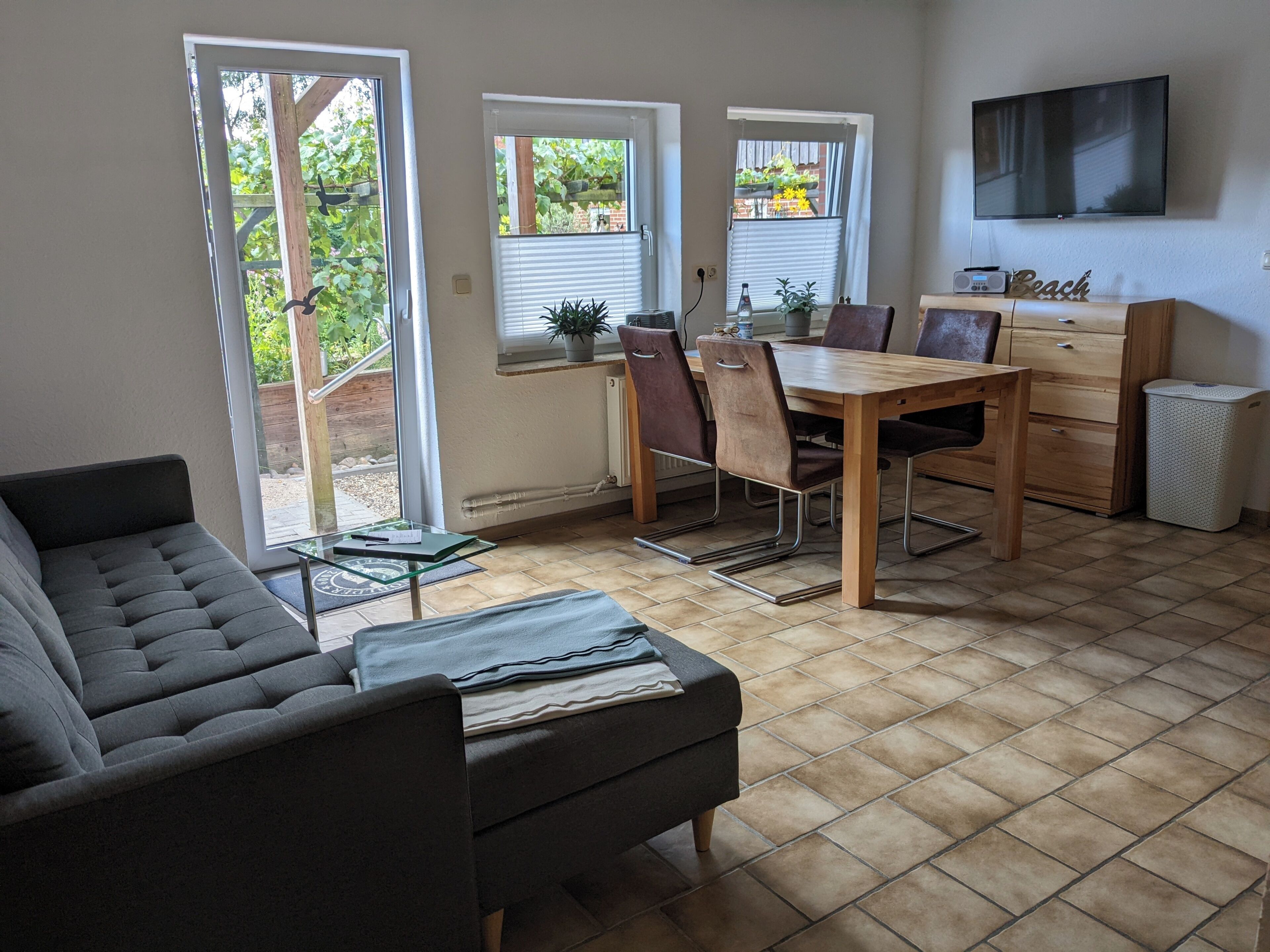 Ein Wohnzimmer mit einer Couch, einem Esstisch mit Stühlen, einem an der Wand montierten Fernseher und einem Fenster mit Jalousien.