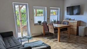 En stue med en sofa, et spisebord med stole, et fjernsyn monteret på væggen og et vindue med persienner.